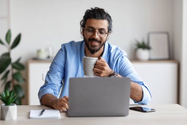 Genç, gülümseyen, serbest çalışan, dizüstü bilgisayar kullanan, evde kahve içen, bilgisayarla uzaktan yakından ilgilenen, uzaktan kumandalı iş fırsatlarının tadını çıkaran, bedava uzay boşluğunun tadını çıkaran Arap bir adam.