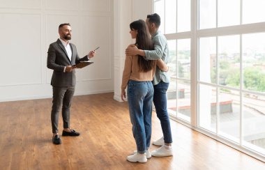 Profesyonel Erkek Emlakçı veya Takım Elbiseli Mimar Milenyum Alıcılarına Yeni Boş Ev Gösteriyor. Eşini kucaklayan koca, eşleri konut binasını ziyaret ediyor, daire seçiyor ve arıyor.