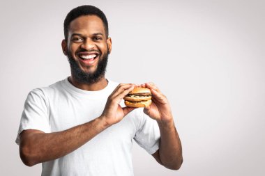 Beyaz arka planda durup kameraya gülümseyen, Burger 'lı Afro-Amerikan Adam Hile Yapıyor. Stüdyo çekimi. Sağlıksız beslenme, abur cubur, aşırı yeme alışkanlığı