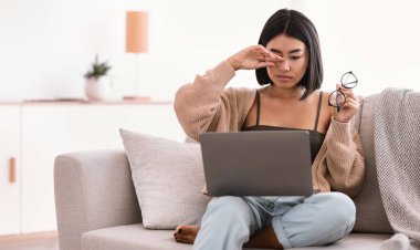 İş yorgunluğu. Uykulu Asyalı kadın göz yorgunluğu çekiyor, gözlük tutuyor, gözleri ovuyor, dizüstü bilgisayarda çalışıyor, oturma odasında kanepede oturuyor. Yaralı gözlere dokunan kadın