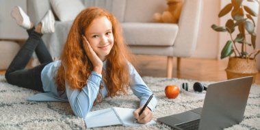 Evde ders çalışıyorum. Neşeli genç kız halıda bilgisayarla yatıyor ve not defterine not alıyor. Liseli kız çevrimiçi kurs izliyor, kameraya bakarak yabancı dil öğreniyor.