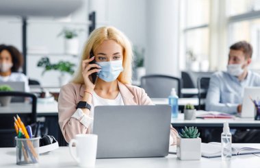 Karantinadan döndükten sonra modern ofiste çalışıyor. Koruyucu maskeli Millennial Lady telefonda konuşuyor ve masada içi antiseptik dolu bir dizüstü bilgisayara bakıyor.