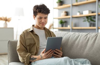 Oturma odasındaki koltukta oturan ve gülümseyen Asyalı adamın dijital tableti tutuş ve kullanış portresi. Mutlu Erkek Sörfleri İnternet Okuma Elektronik Kitap Okuma, Hafta sonu Boş Zaman Geçirmenin Keyfi