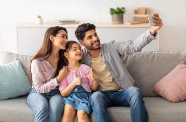 Aile özçekimi. Mutlu Arap adam karısı ve küçük kızıyla fotoğraf çekiyor, evdeki kanepede oturuyor, hafta sonları birlikte vakit geçiriyorlar. Bağlanma ve yaşam tarzı kavramı