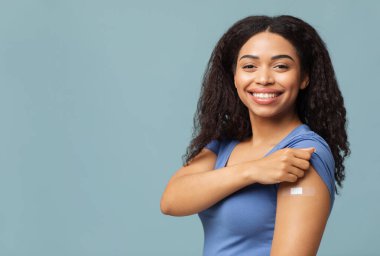 Covid-19 aşımı yaptırdım. Mutlu Afro-Amerikalı kadın aşı yaptırdıktan sonra kolunu gösteriyor ve kameraya gülümsüyor, mavi stüdyo arka planında poz veriyor. Coronavirus aşısı