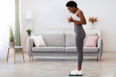 Zayıflamada başarılı. Neşeli siyahi kadın ağırlığını evde tartıyor ve sevinçle yumruklarını sallıyor. Büyük Kilo Verme Konsepti. Metin için Tam Uzunluk, Yan Görünüm, Boş Alan