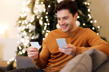 Online Shopping Concept. Portrait Of Smiling Male Buyer Using Cell Phone, Holding And Looking At Debit Credit Card Purchasing Things On Internet Shop Sitting On Couch At Home In Living Room