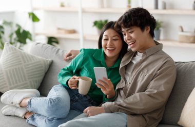 Akıllı telefon kullanan ve evdeki kanepede dinlenirken kahve içen genç Asyalı pozitif bir çift. Mobil cihazda mutlu bin yıllık aile web tarayıcıları ve sıcak içeceğin keyfini çıkarın.