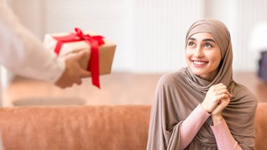 Müslüman Kadın, evdeki koltukta oturan adamdan paketlenmiş hediye paketi alıyor. Teslimat, doğum günü ve yeni yıl kutlaması konseptini sunar. Kırpılmış, Seçici Odaklanma