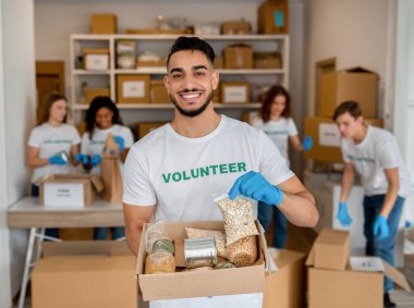 Genç Arap gönüllü hayır kurumunda çalışıyor, yiyecek bağış kutusunu tutuyor ve kameraya gülümsüyor. Hayır kurumunda kameraya poz veren adam.