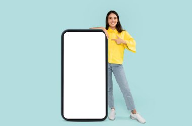 Cool cheerful stylish pretty young brunette woman pointing at huge phone with white blank mockup screen, showing nice online offer or recommending mobile app, blue background, copy space