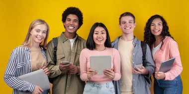 Uzaktan eğitim için modern dijital cihazlar. Sırt çantalı, telefonlu, tabletli ve dizüstü bilgisayarlı mutlu genç insanlar kameraya bakar, turuncu arka planda izole edilmiş, stüdyo çekimi, boş alan