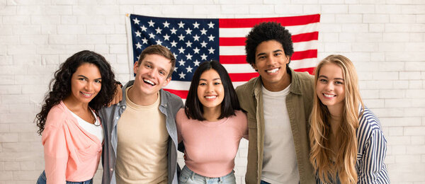 Fun education, language courses, learn english and exchange students. Young smiling people of different nationalities hug in college with large USA flag on white brick wall, free space, studio shot