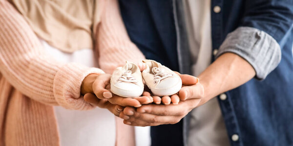 Closeup shot of expectant muslim family holding small baby shoes in their hands, unrecognizable pregnant islamic lady in hijab and her loving husband enjoying future parenthood, cropped image