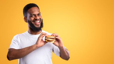 Abur cubur. Sarı Stüdyo Arkaplanda Burger Poz Veren Mutlu Afrikalı Amerikalı Adam, Kameraya Gülümsüyor. Beslenme ve aşırı yeme alışkanlığı, Fast Food Aşığı Konsepti