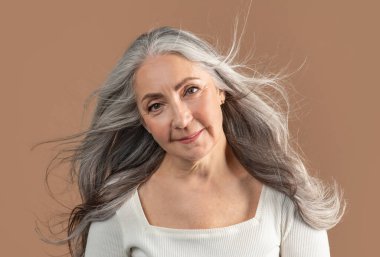 Kahverengi stüdyo arka planında uzun ve sağlıklı gri uçan saçlarla poz veren gülümseyen yaşlı kadın portresi. Güzel olgun bir bayan kameraya poz veriyor ve gülümsüyor. Saç bakım ve moda konsepti