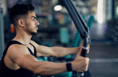 Spor salonunda güvertede çalışan kendinden emin bir Arap sporcu, spor kulübünde modern ekipman kullanan, kol ve sırt kasları üzerinde çalışan, yan görünümü olan motive olmuş orta doğulu vücut geliştiricinin portresi.