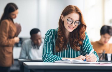 Eğitim, Lise, Öğrenme ve İnsanlar Konsepti. Gözlüklü güzel bir kız öğrencinin portresi. Sınıfta yazı sınavı. Sınıf arkadaşlarıyla aynı masada oturuyor.