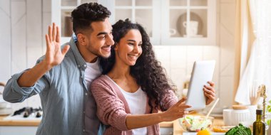 Modern Müslüman çift evdeki mutfakta dijital tabletle video görüşmesi yapıyor, neşeli Arap eşleri kameraya el sallıyor, birine selam veriyor, arkadaşlarına ya da ailesine selam veriyor, boş bir alana el sallıyor.