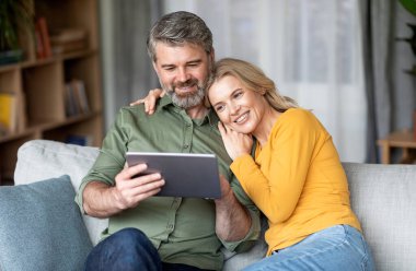 Boş Zaman Teknolojileri. Mutlu Orta Çağ Eşleri Evde Dijital Tablet Kullanıyor, Gülümseyen Olgun Eş ve Koca Modern Gadget ve Kucaklayan, Keyifli Ev Eğlencesi