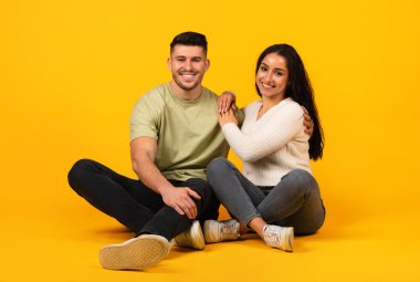 Cheerful millennial arab guy and lady in casual sit on floor, enjoy date, isolated on orange background, studio. Couple relax in free time, love, relationship, advice and recommendation, ad and offer