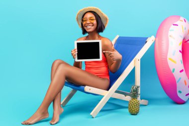 Mavi arka planda şezlongda otururken, bikini giymiş mutlu siyah kadın tableti boş ekranla göstererek tasarım için maket yapıyor. Uygulama ya da web sitesi için boşluk gösteren genç bayan