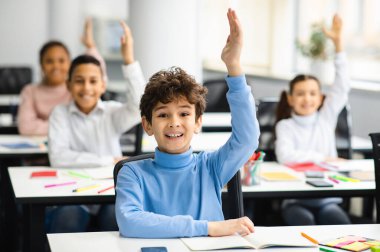Eğitim, Ortaokul, Öğrenme ve İnsanlar Konsepti. Mutlu okul çocuğu cevap için elini kaldırıyor, sınıfta çeşitli gülümseyen sınıf arkadaşlarıyla oturuyor, zevkle çalışıyor.