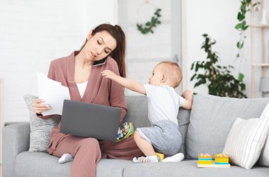 İş ve Annelik. Sinirli bir anne, bebeğinin dikkatini dağıtırken evde çalışmaya çalışıyor, annelik stresi çeken genç bir kadın, boş alan.