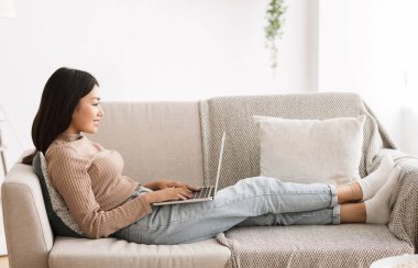 Online eğitim. Dizüstü bilgisayar kullanan Asyalı bir kız. Evdeki rahat koltukta oturuyor.