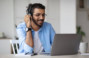Gülümseyen Hintli genç adam dizüstü bilgisayarında kulaklık ve gözlük takıyor, online eğitimin ve uzaktan öğrenmenin tadını çıkarıyor, evde oturup bilgisayar ekranına bakıyor, boş alan arıyor,