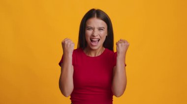 Kafkasyalı genç bir kadın şaşkın bir ifadeyle ayakta duruyor. Koyu renk saçları, parlak sarı arka planla çelişiyor. Dikkat çeken çarpıcı bir görüntü oluşturuyor..