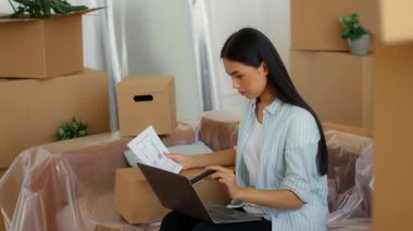 Genç bir Asyalı kadın, yeni boşaltılmış bir dairede kutuların arasında oturmuş, dizüstü bilgisayar kullanarak, ailesinin yeni evine taşınmasına hazırlanırken belgeleri düzenliyor..