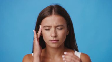 Uzun saçlı beyaz bir kadın acı dolu bir ifadeyle gösteriliyor, sanki migren ya da baş ağrısı gibi şakağına hafifçe dokunuyor. Canlı mavi arka plan onun odaklanmış davranışını arttırıyor..