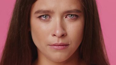 Genç beyaz bir kadın, gözlerinde yaşlarla derin bir duygusal ifade sergiliyor. Canlı pembe arka plan duygularını vurguluyor, çarpıcı bir görsel etki yaratıyor..