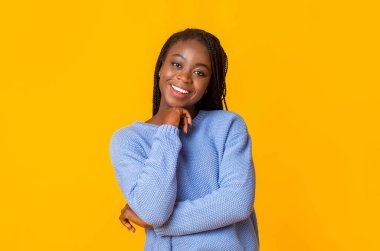 Sarı stüdyo arka planında gülümseyen mavi kazaklı afro kadın.