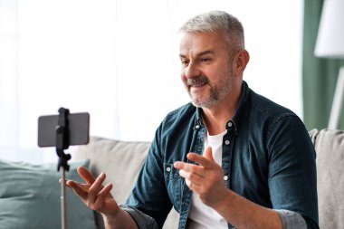 Çekici olgun adam vlogger ya da blogger evden yayın yapıyor, rahat oturma odasında koltukta oturuyor ve cep telefonuyla video çekiyor, tripoda yerleşti, kıdemli adam görüntülü konuşma yapıyor, boşluğu kopyalıyor.