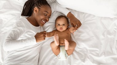 Happy Maternity. Above top high angle view portrait of positive African American woman lying in bed and playing with her cute little baby, touching infants arms, spending time on white bedsheets