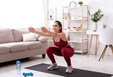 Evde kal, formda kal. Spor kıyafetli güzel bir kız dairesinde çömeliyor.