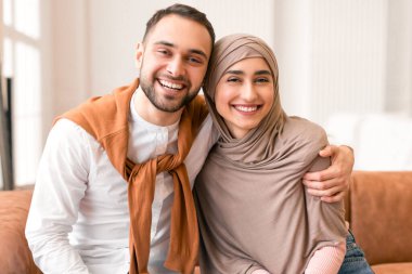 Mutlu evlilikler. Neşeli Müslüman eşler kameraya gülümsüyor ve evdeki koltukta oturuyorlar. Hijab 'da karısını kucaklayan sevecen bir Arap koca. Aile, Romantik İlişki Konsepti