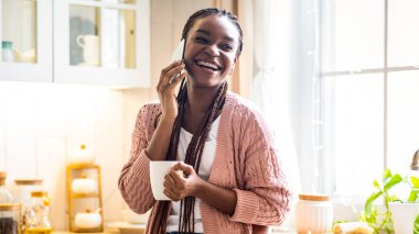 Cep telefonuyla konuşan ve mutfakta kahve içen neşeli siyahi kadının portresi, evde dinlenirken keyifli mobil sohbetin tadını çıkaran Afro-Amerikan kadını,