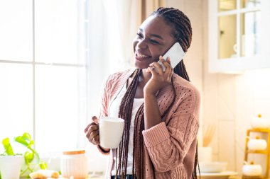 Neşeli siyah kadın kahve içiyor ve mutfakta telefon görüşmesi yapıyor. Genç Afrikalı Amerikalı bayan sıcak içecekle bardağı tutuyor ve keyifli mobil bir sohbetin tadını çıkarıyor,