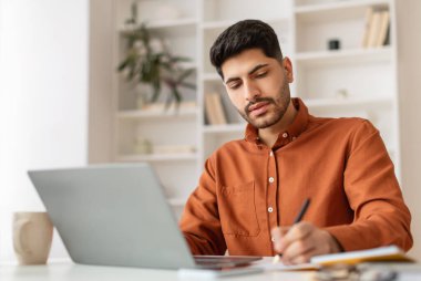 Odaklanmış Orta Doğulu genç bir adam dizüstü bilgisayarla çalışıyor, not alıyor, milenyum erkeğiyle meşgul, internetten ders çalışıyor, bilgisayar kullanıyor ve kontrol listesi yazıyor.
