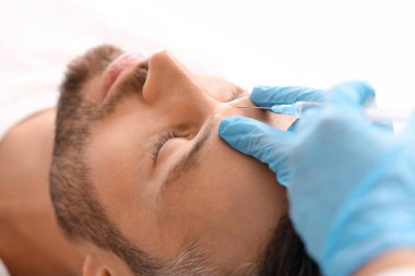 Klinikte güzellik iğnesi yaptıran sakallı adama yakın çekim. Kozmetik uzmanı, yakışıklı bir adam için yüz doldurma işlemi için mavi lastik eldivenleri teslim etti. Yüz bakımı, erkek kozmetoloji konsepti.