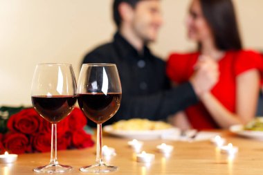 Romantic Date. Selective focus on two glasses of red wine on the table in the foreground. Lovely couple spending time together at home or cafe, having dinner, holding hands in blurred background