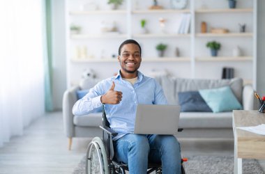 Dizüstü bilgisayarı olan mutlu, özürlü siyahi bir adamın portresi başparmağını kaldırıyor, içerde kameraya gülümsüyor. Tekerlekli sandalyedeki belden aşağısı felçli genç adam covid salgını sırasında online iş öneriyor.