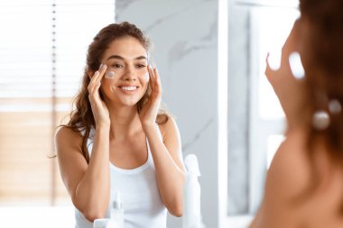 Genç bir kadın evde modern banyoda aynaya bakarken yüzüne ve yanaklarına nemlendirici krem sürüyor. Cilt Bakımı ve Tedavi Konsepti. Seçici Odaklanma