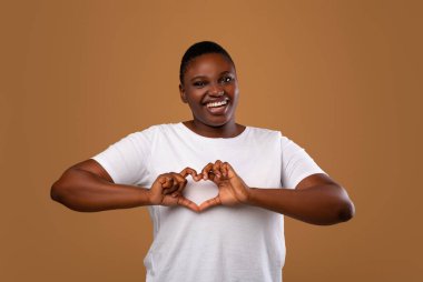 Nazik, tombul, kalp şeklinde elleri olan, göğsünün yanında aşk jestleri yapan, şefkat ve kendini beğenmiş, kameraya gülümseyen, kahverengi stüdyo duvarının başında duran,