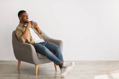 Rahatlamış siyah adam, sabah kahvesinin keyfini çıkarıyor gri koltukta oturmuş, içerideki boşluğu kopyalıyor. Minimalizm yaşam tarzının rahatlığı. Hafta sonu tatili konsepti. Reklam Afişi
