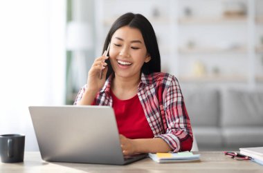 Telekomünikasyon konsepti. Neşeli Asyalı kadın cep telefonuyla konuşuyor ve evde dizüstü bilgisayarla çalışıyor, neşeli Koreli kadın bilgisayar kullanıyor ve keyifli cep telefonu konuşmaları yapıyor.