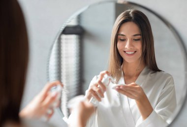 Güzellik rutini. Çekici, genç bir kadın banyoda aynanın yanında dururken pamuk pedin üzerine makyaj sütü sürüyor, Gülümseyen, Güzel Bayan Temizleme Cildi, Evde Günlük Skincare Yapıyor